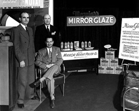 Une image en noir et blanc de trois hommes en complet à côté d’un présentoir de produits et de panneaux Meguiar’s.